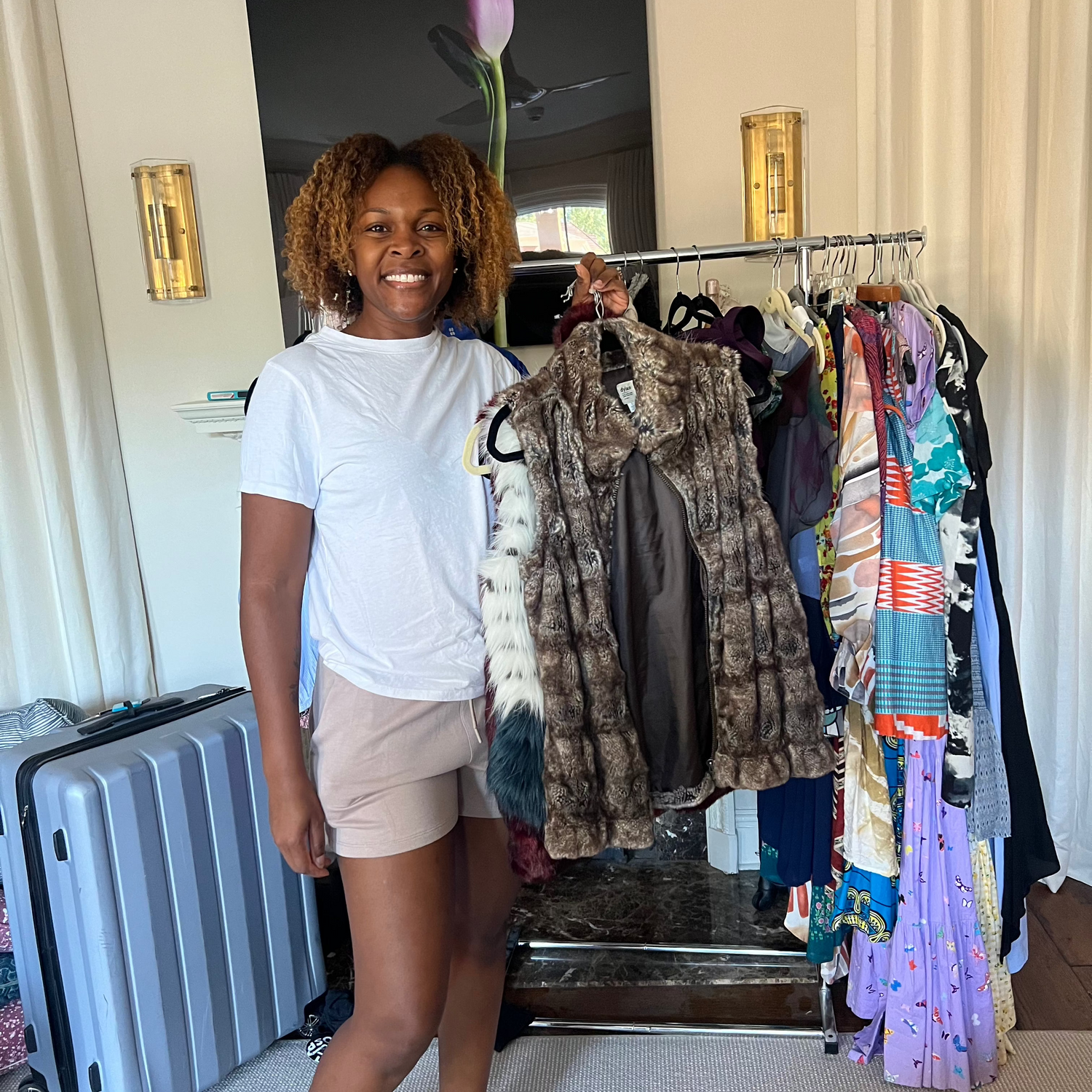 Woman standing in a room with a rack of clothes and a suitcase.